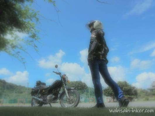 鶴とイチョウ柄の黒系アロハシャツを着た女性が、YAMAHA SR125の前で空を見上げるように立っている。芝生と青空、夏の終わりのお散歩ツーリング風景