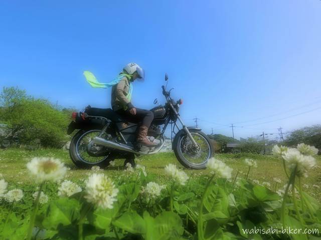 シロツメクサ咲く草地に停めたSR125に跨るバイク乗り。風でなびく緑のスカーフが薫風の季節を象徴するセルフショット