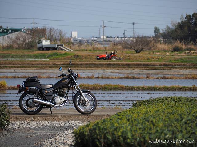 田植え前、水だけが張られた田んぼの脇に佇むYAMAHA SR125。奥には代掻き中の赤いトラクターが見える、初夏直前の農村風景