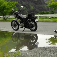 曇天の下、水たまりにSR125が映る。バイクに向かって歩くライダーの足元も写り、梅雨入り前の気配を感じる5月の風景