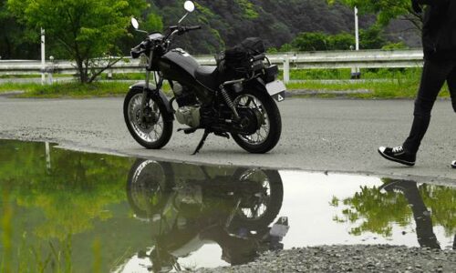 曇天の下、水たまりにSR125が映る。バイクに向かって歩くライダーの足元も写り、梅雨入り前の気配を感じる5月の風景