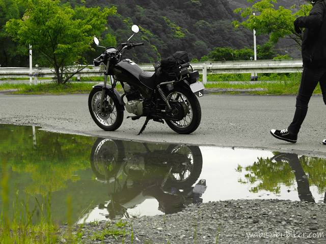 曇天の下、水たまりにSR125が映る。バイクに向かって歩くライダーの足元も写り、梅雨入り前の気配を感じる5月の風景