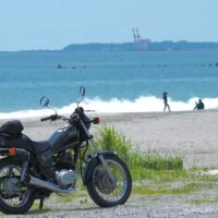 海辺に停めたYAMAHA SR125の奥に、白波が寄せる浜辺と写真を撮り合うカップルのシルエットが写っている。空には雲が浮かび、湾岸のクレーンが遠くにぼんやり見える。バイクと夏の海、そして他者の記録が静かに共存する一枚