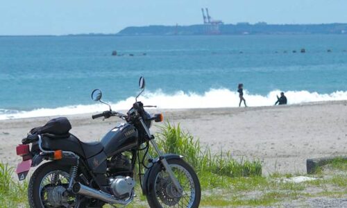 海辺に停めたYAMAHA SR125の奥に、白波が寄せる浜辺と写真を撮り合うカップルのシルエットが写っている。空には雲が浮かび、湾岸のクレーンが遠くにぼんやり見える。バイクと夏の海、そして他者の記録が静かに共存する一枚