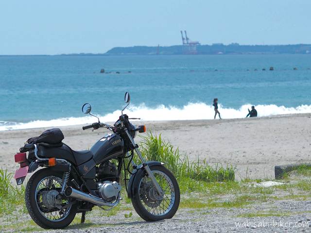 海辺に停めたYAMAHA SR125の奥に、白波が寄せる浜辺と写真を撮り合うカップルのシルエットが写っている。空には雲が浮かび、湾岸のクレーンが遠くにぼんやり見える。バイクと夏の海、そして他者の記録が静かに共存する一枚