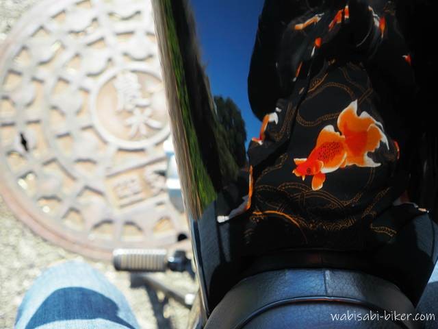 バイクの黒いタンクに、金魚柄のアロハシャツが鮮やかに映り込んでいる。 足元には「農水」と刻まれたマンホール蓋があり、田舎道を連想させる一枚