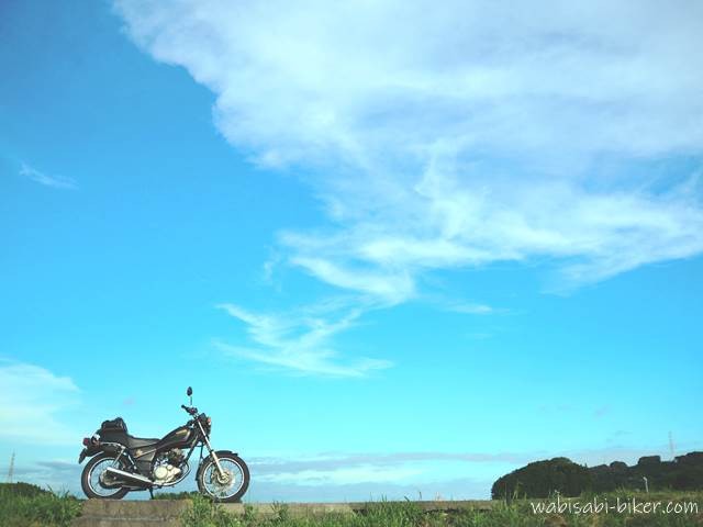 広い夏の青空を背景に、道路脇に停まるバイク(YAMAHA SR125)が小さく写っている。雲の形と空の青さが印象的な、シンプルな構図