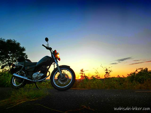 夕暮れの空を背景に停められた黒いバイク(YAMAHA SR125)。舗装路の上に立ち、薄明るい空と草のシルエットに囲まれながら、ライトの光がかすかに灯る
