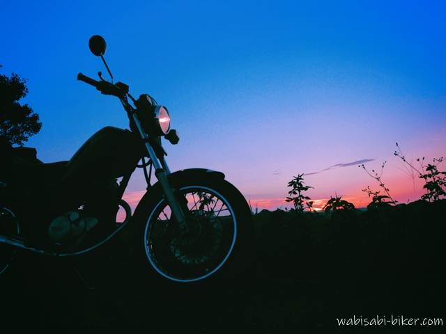 夕暮れ空を背景にシルエットで浮かび上がる黒いバイク（YAMAHA SR125）。フロントライトが灯り、空は青から紫、そして橙に移ろうグラデーションを描いている。