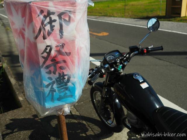 「御祭禮」と書かれた赤と青の提灯がビニールで覆われ、道路脇の支柱に取り付けられている。背景には黒いバイク（YAMAHA SR125）が停車しており、祭りの準備が進む町の一角を切り取った風景。