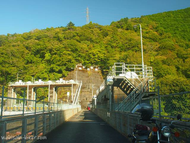 笹間川ダムの天端から撮影された写真。手前右に黒いバイク（YAMAHA SR125）の一部が写り込み、奥にはダム施設の構造物と山の斜面が見える。秋の光に照らされ、緑がやや色づき始めている風景。