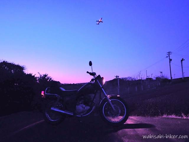 夕暮れから紫に染まる空の下、小道の脇に停めたバイクSR125のシルエットと、上空を離陸していく飛行機をセルフタイマー撮影で捉えた写真。