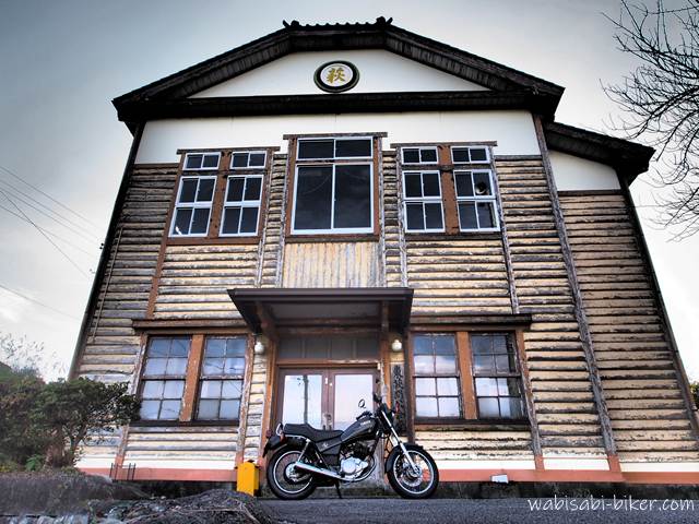 茶畑に建つ木造のレトロ建築「東萩間公会堂」を正面から捉え、その前に納車間もないバイクSR125を停めた写真。板張りの外壁と古びた窓枠が昭和初期の面影を残し、曇り空の下で静かに佇んでいる。走り慣れた道なのに存在に気づかず、興奮しながらバイクと並べて撮った一枚。