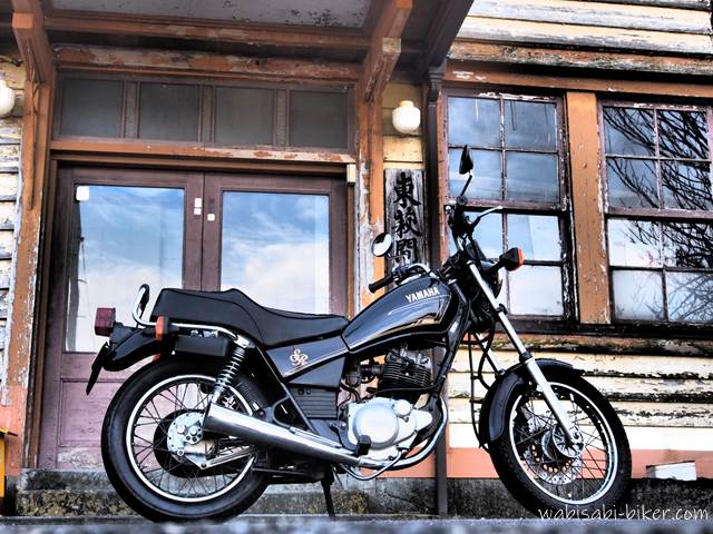 古びた公会堂の窓と外壁を背景に、バイクSR125を横から大きく配置した写真。窓ガラスには冬空と枯れ木が映り込み、木製の窓枠や塗装の剥がれた外壁に昭和初期の年月が滲んでいる。古さが凝縮された窓周辺に興奮してバイクを寄せて撮った一枚。