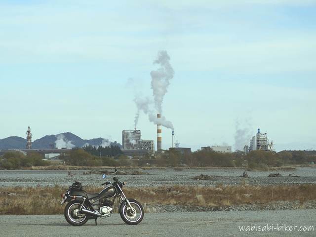 大井川の河川敷に停めた黒いバイクSR125を手前に据え、奥にパルプ工場の煙突と施設群を配置した写真。冬の無風の日で、煙突の煙がまっすぐ上に伸びている。川の水位が低く、乾いた河原の質感が広がる、冬らしさを記録した一枚。