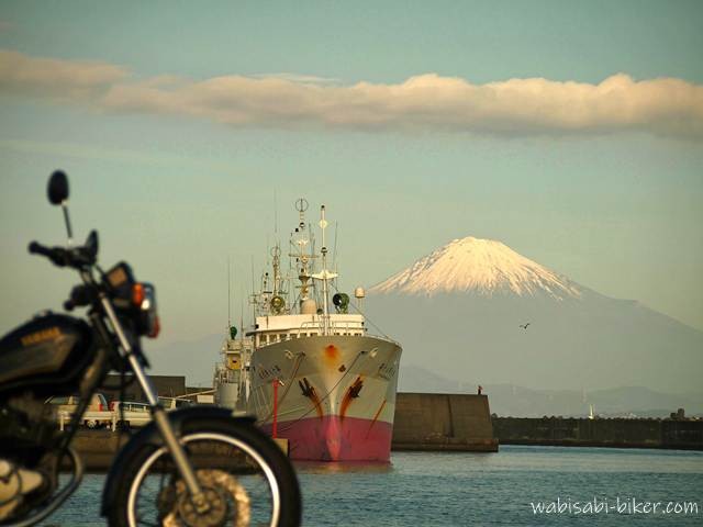 港に停泊する漁船の奥に雪化粧した富士山を主役として捉え、左端に黒いバイクSR125をぼかして控えめに配置した写真。青い海と冬の澄んだ空気の中で、富士山の白が静かに際立つ構図に整えた一枚。