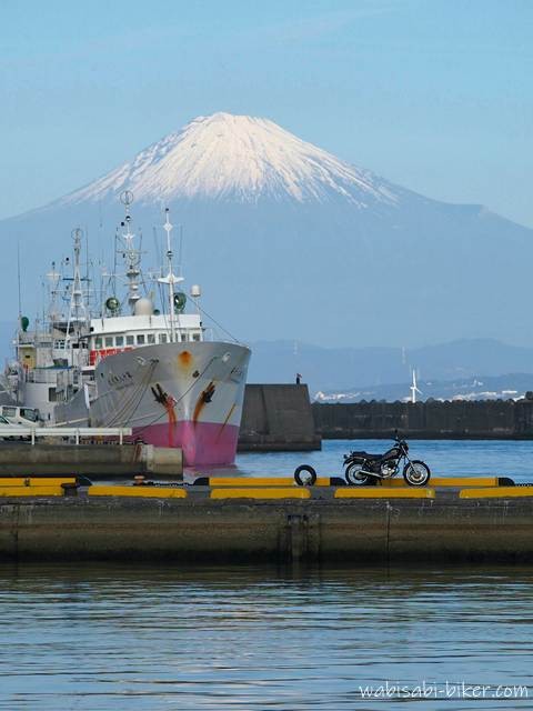 雪化粧した富士山と停泊する漁船を大きく捉え、その手前の岸壁に黒いバイクSR125を小さく配置した写真。望遠レンズで圧縮し、バイクの黒が奥の防波堤の黒壁に埋もれない位置を選びながら、風景の中でバイクの小ささを際立たせた一枚。