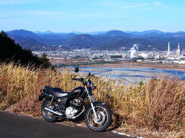 冬枯れの草が揺れる高台の小道に停めた黒いバイクSR125。眼下には大井川と町並み、遠くの山並みまでが澄んだ冬空に広がる。納車後初めて迎えた年明けに、冬の空気ごとバイクを残したくて撮影した一枚。