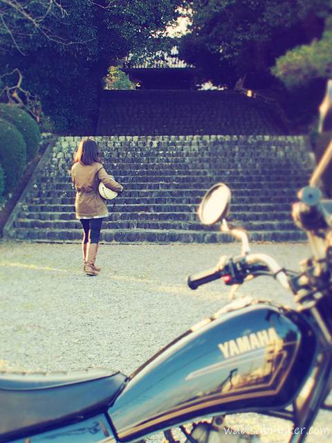 お寺の参道と、石段に向かうライダーの後ろ姿を捉えたセルフスナップ。手前に黒いバイクSR125のタンクとミラー越しに、ヘルメットを抱えたバイク乗りを写した一枚。