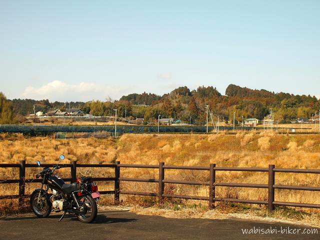 冬枯れの草地を見渡す柵の前に停めた黒いバイクSR125。黄金色の枯れ草が広がる空き地の向こうに、遠くの集落と冬空が続く。納車まもない頃、冬の乾いた空気ごと残したくて撮影した一枚。