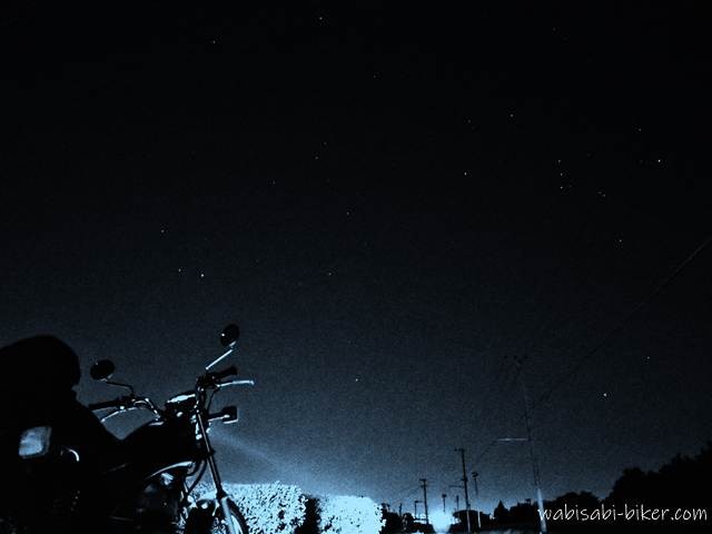 夜の道路沿いに停めたバイクSR125のシルエットと、ライトに照らされた茶畑の先に続く道。その上の夜空に冬の大三角がかすかに浮かんでいる。