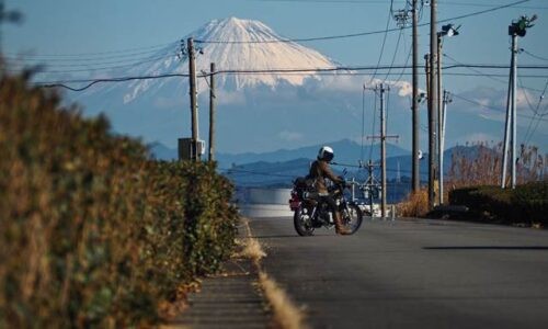 遠くに大きくそびえる富士山を主役に、郊外の道路でSR125に乗ったバイカーが向きを変えながらカメラを確認する一瞬が、景色の中の小さな存在として写っている。