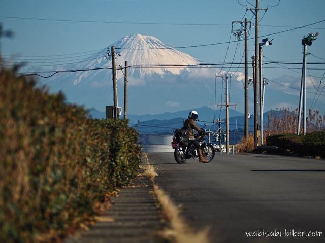 遠くに大きくそびえる富士山を主役に、郊外の道路でSR125に乗ったバイカーが向きを変えながらカメラを確認する一瞬が、景色の中の小さな存在として写っている。