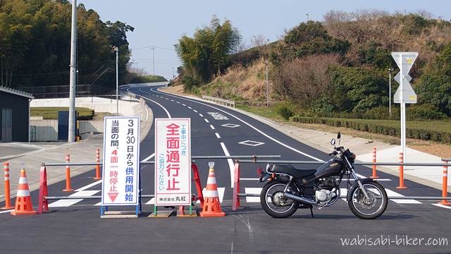 開通前で通行止めとなっている新しい道路の手前に、規制コーンと「全面通行止」「3月30日から供用開始」と書かれた看板が並ぶ風景。右側に黒いバイクSR125を停め、奥へ伸びる道路と周囲の茶畑や斜面を写した一枚。