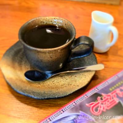 陶器のカップに注がれたホットコーヒーとミルクポット、手前に漫画が写ったテーブルの上の一枚。喫茶店での休憩シーン