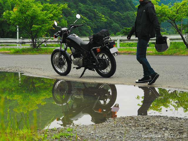 水たまりにリフレクションが映る黒いバイクと、ヘルメットを持って歩く人物。曇り空の下、濡れた地面と新緑が印象的な風景