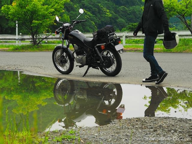 水たまりにリフレクションが映る黒いバイクと、ヘルメットを持って歩く人物。曇り空の下、濡れた地面と新緑が印象的な風景