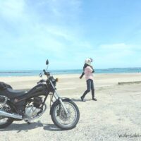 初夏の海辺に停めたSR125。奥にはピンクのリネンシャツを着た人物が歩いている。バイクとの距離感が、静かな夏の入口を感じさせる。
