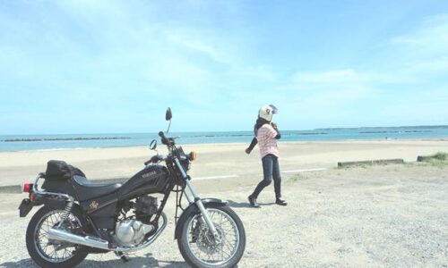 初夏の海辺に停めたSR125。奥にはピンクのリネンシャツを着た人物が歩いている。バイクとの距離感が、静かな夏の入口を感じさせる。