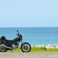 海を背景に停められたYAMAHA SR125。水平線まで広がる青い海と、雲の浮かぶ夏空がバイクを引き立てている。人物は写っておらず、静かな浜辺に佇むバイクだけが画面の主役となっている