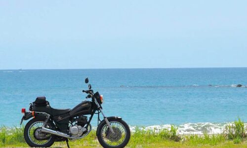 海を背景に停められたYAMAHA SR125。水平線まで広がる青い海と、雲の浮かぶ夏空がバイクを引き立てている。人物は写っておらず、静かな浜辺に佇むバイクだけが画面の主役となっている
