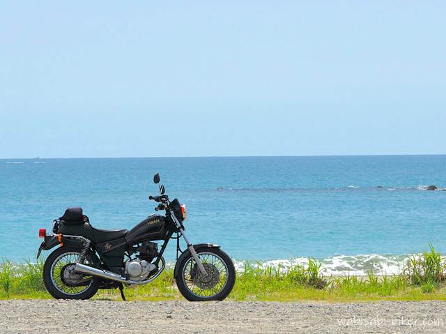 海を背景に停められたYAMAHA SR125。水平線まで広がる青い海と、雲の浮かぶ夏空がバイクを引き立てている。人物は写っておらず、静かな浜辺に佇むバイクだけが画面の主役となっている