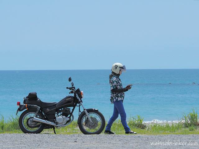 海辺を花柄アロハシャツとヘルメット姿のライダーがスマホを手に歩いている。バイク（YAMAHA SR125）は手前に停車しており、背景には青い海が広がる。夏の空気を感じさせる、自然な動きのある一枚