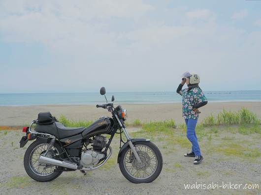 海辺に停めたYAMAHA SR125の隣で、花柄アロハシャツを着たライダーがヘルメット姿で立っている。シャツは「劇場版 呪術廻戦 0」の夏油傑のアロハ。背景には穏やかな海と空が広がり、夏らしさと落ち着きが同居する一枚