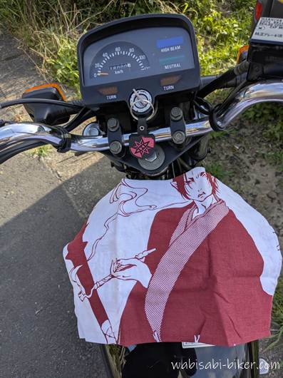 バイクのハンドル越しに見た計器盤とタンク。その上に赤と白の手拭いが広げられ、アニメ「鬼灯の冷徹」のキャラクター鬼灯が描かれている。お盆休みの記録として、魔除けを兼ねて持参した一枚