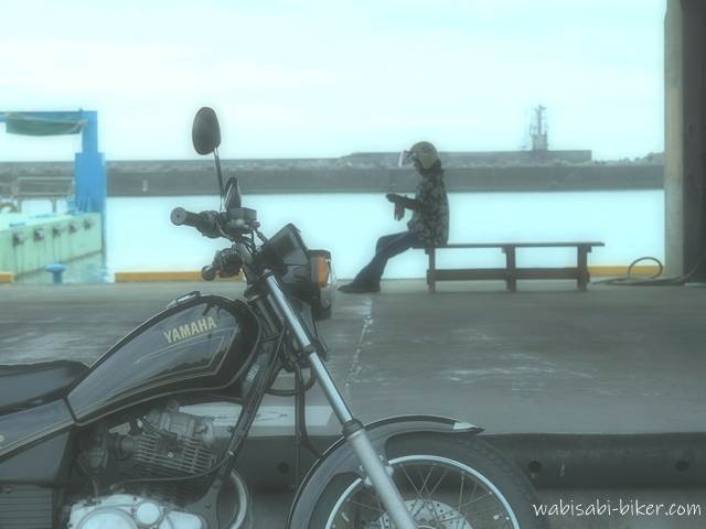 夏の漁港の荷捌き場。手前には黒いバイク(YAMAHA SR125)、奥のベンチにヘルメット姿のライダーが座り、静かな海を前に休憩している。お盆休みで人影はなく、淡い光が港内に満ちている