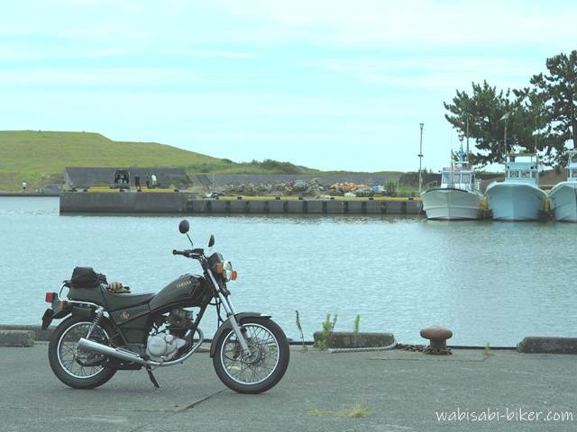 漁港の岸壁に停めた黒いバイク(YAMAHA SR125)。背後には停泊する数隻の漁船と、堤防越しに見える緑の斜面。対岸には数人の人影があり、夏休み中の港らしい穏やかな空気が漂っている