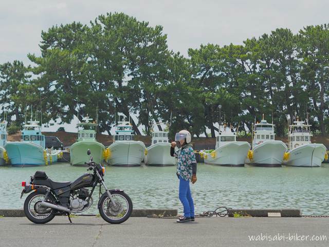 漁船が横一列に並ぶ港の岸壁。前景に黒いバイク(YAMAHA SR125)が停まり、その奥で花柄シャツと青いパンツを着たライダーが立ち、片腕を上げている。曇り空の下で撮影され、服装の全体像を記録として残した一枚