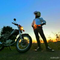 バイク(YAMAHA SR125)の横に立つ女性ライダー。白いワイシャツをズボンにインし、黒パンツと黒いスニーカーを合わせている。夕焼けを背景に腰に手を当てたポーズで立っており、気取りながらもどこか滑稽な雰囲気