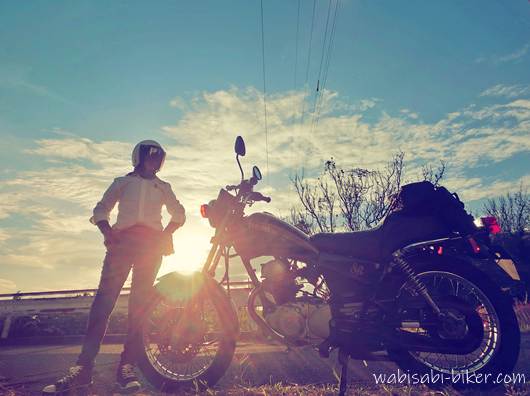 夕陽を背に、黒いバイク（YAMAHA SR125）の横に白いワイシャツ姿のライダーが立っている