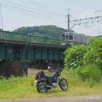 草地に停めた黒いバイク(YAMAHA SR125)の奥を、鉄橋を走る電車が通過している。橋梁は低く、古びたレンガの橋脚が特徴。背景には緑の山々が広がり、静かな初夏の風景が広がる。