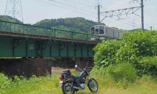草地に停めた黒いバイク(YAMAHA SR125)の奥を、鉄橋を走る電車が通過している。橋梁は低く、古びたレンガの橋脚が特徴。背景には緑の山々が広がり、静かな初夏の風景が広がる。
