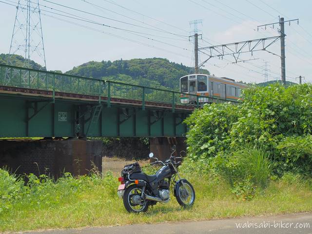 草地に停めた黒いバイク(YAMAHA SR125)の奥を、鉄橋を走る電車が通過している。橋梁は低く、古びたレンガの橋脚が特徴。背景には緑の山々が広がり、静かな初夏の風景が広がる。