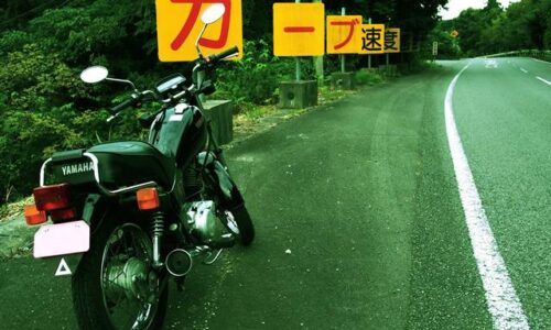 道路脇に停められた黒いバイク(YAMAHA SR125)が写っている。左奥には「カーブ速度」と書かれた警告看板が立ち並び、右側に緩やかなカーブの道路が続いている。看板の文字が強調され、構図にユーモアと緊張感が漂う一枚。