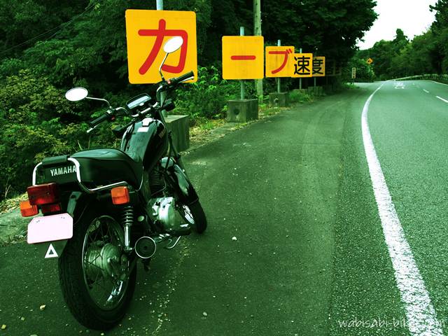 道路脇に停められた黒いバイク(YAMAHA SR125)が写っている。左奥には「カーブ速度」と書かれた警告看板が立ち並び、右側に緩やかなカーブの道路が続いている。看板の文字が強調され、構図にユーモアと緊張感が漂う一枚。