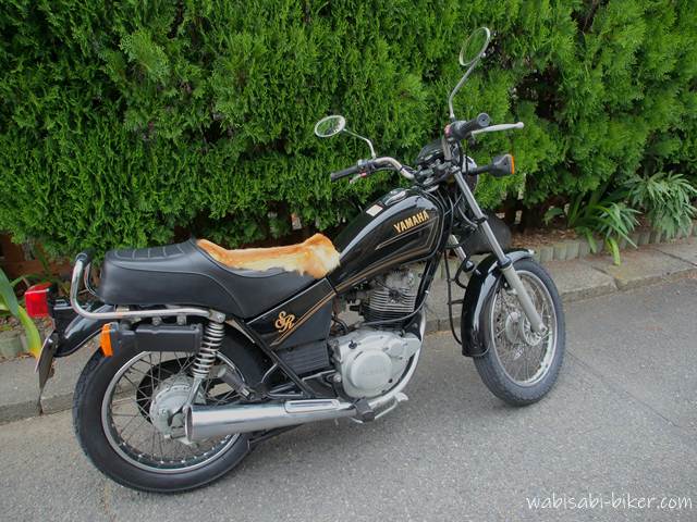 黒いバイク(YAMAHA SR125)のシート上に鹿の毛皮が置かれている。生垣のある路上で停車中の様子。異質な毛皮が目を引く1枚